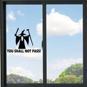 May include: A black vinyl decal applied to a glass window pane. The decal features a silhouette of a wizard wearing a pointed hat, holding a sword in one hand and a staff in the other, with the text YOU SHALL NOT PASS! printed below it.