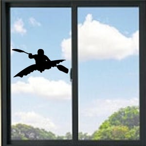 Op de afbeelding: Zwart silhouet van een kajakker die op het water peddelt, gezien door een raam. De kajakker zit in een kajak met een peddel. De achtergrond toont een blauwe lucht met witte wolken en groene bomen.