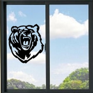 May include: A black vinyl decal featuring the stylised head of a roaring bear with an open mouth and sharp teeth. The graphic design uses bold lines and solid black shapes to create a high-contrast silhouette applied to a glass window pane.
