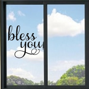 May include: A black vinyl decal applied to a glass window pane. The decal features the text "bless you" written in a flowing, cursive script font. The lettering is positioned on the left side of the window, with a clear view of the sky.