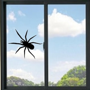 May include: A black spider silhouette decal applied to a glass window pane. The spider graphic features a central body with eight long, pointed legs extending outward in a crawling position, creating a high-contrast visual against the clear glass.