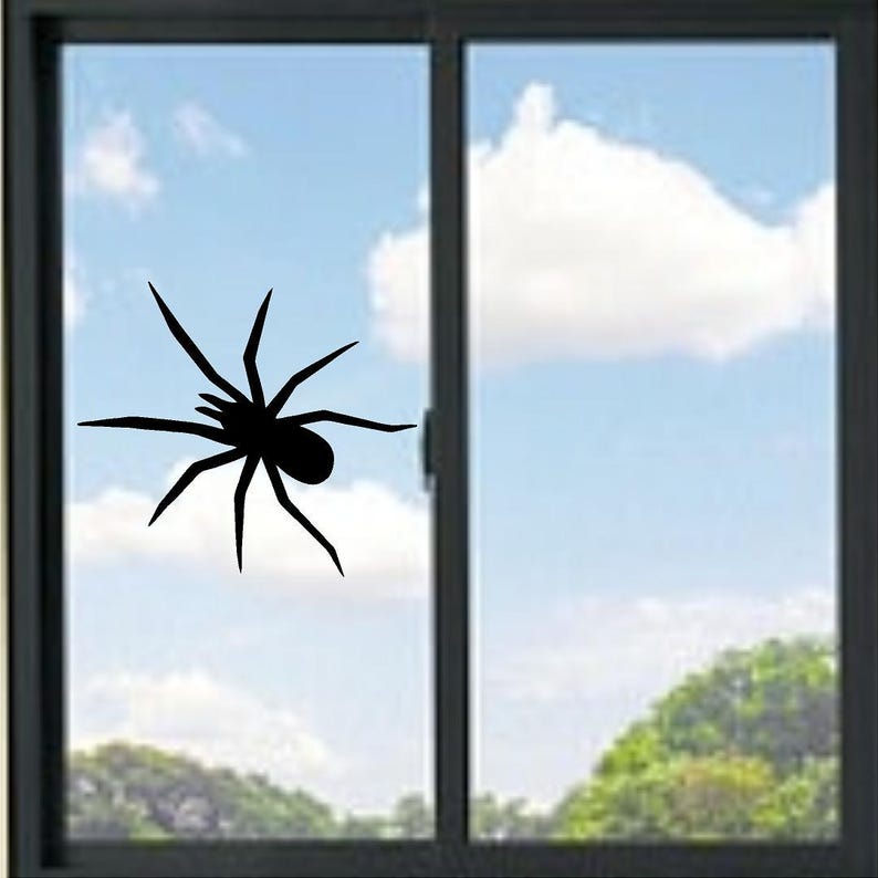 May include: A black spider silhouette decal applied to a glass window pane. The spider graphic features a central body with eight long, pointed legs extending outward in a crawling position, creating a high-contrast visual against the clear glass.