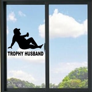 Pode incluir: Um autocolante de vinil preto aplicado numa janela de vidro, apresentando a silhueta de uma pessoa com um boné, reclinada enquanto segura uma bebida, acompanhada pelo texto preto em maiúsculas que diz TROPHY HUSBAND sob a figura.