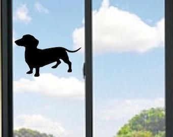 May include: A black silhouette of a dachshund dog is affixed to a window. The dog is in profile, with its body and legs clearly defined. The background shows a blue sky with white clouds and green trees.