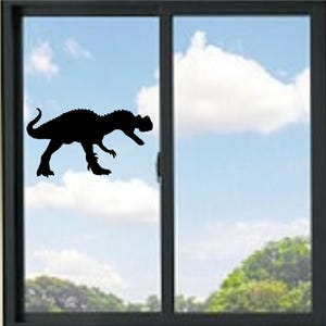 May include: A black silhouette of a dinosaur decal applied to a glass window pane. The dinosaur is depicted in a side profile, standing on its hind legs with its tail curved upwards and its mouth open, showing sharp teeth and a jagged back ridge.