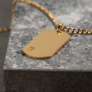 May include: A gold chain necklace with a gold dog tag pendant. The dog tag has a small inscription on it.