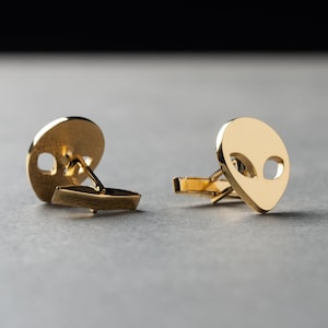 May include: A pair of gold-toned metal cufflinks, each featuring a polished, flat, alien head-shaped face with two cut-out oval eye sockets. The cufflinks have a standard hinged toggle mechanism on the back for securing through shirt cuffs.