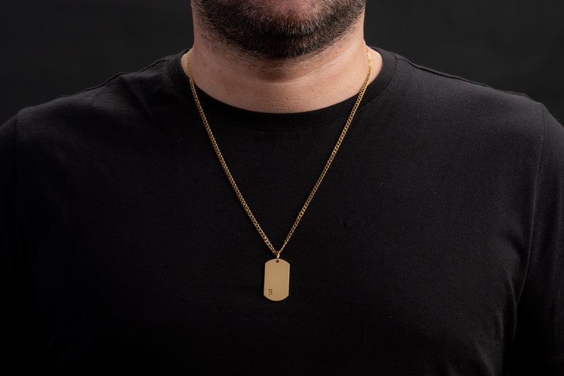 May include: A gold chain necklace with a gold dog tag pendant. The dog tag has the letter "H" engraved on it.