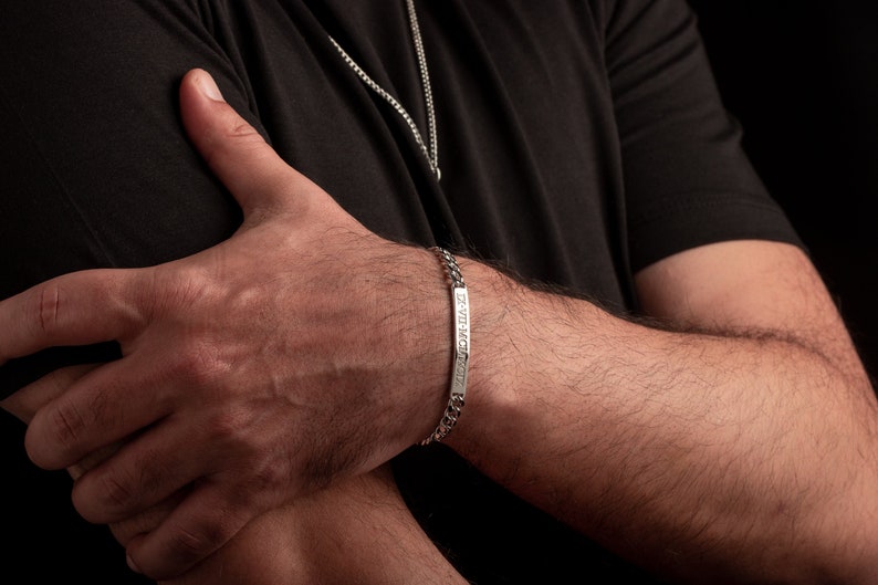 K&ouml;nnte beinhalten: Ein silbernes Kettenarmband mit einem rechteckigen Anh&auml;nger, auf dem "De Vita Scio" steht.