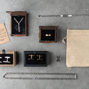 May include: A set of men's jewelry in various boxes and a burlap bag. The jewelry includes a silver chain necklace with a rectangular pendant, a silver chain bracelet, a pair of silver cufflinks, a gold ring, and a silver chain.