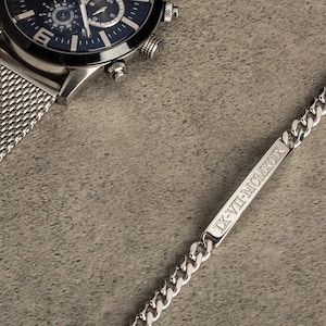 K&ouml;nnte beinhalten: Eine silberne Uhr mit schwarzem Zifferblatt und einem Milanaise-Armband sowie ein silbernes Kettenarmband mit einem rechteckigen Anh&auml;nger, auf dem der Text "IX-VII-MMCMXCIX" eingraviert ist.