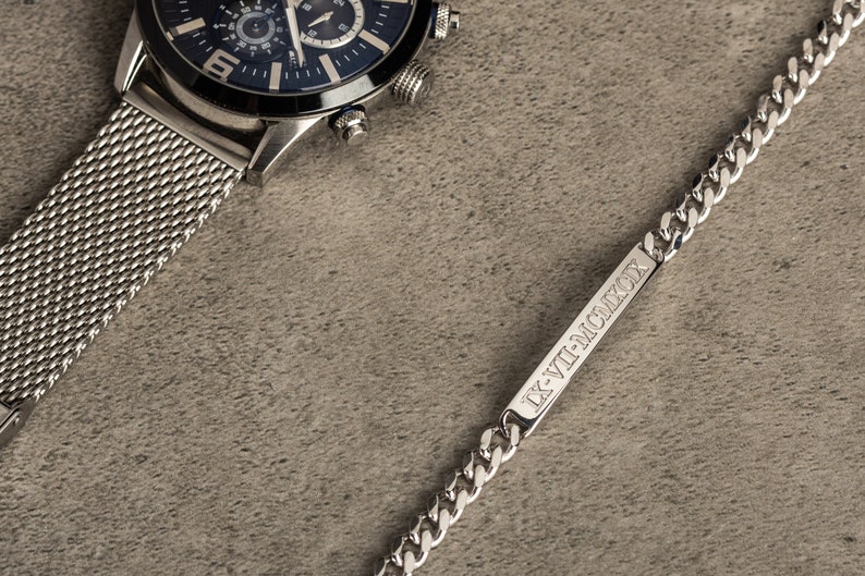 K&ouml;nnte beinhalten: Eine silberne Uhr mit schwarzem Zifferblatt und einem Milanaise-Armband sowie ein silbernes Kettenarmband mit einem rechteckigen Anh&auml;nger, auf dem der Text "IX-VII-MMCMXCIX" eingraviert ist.