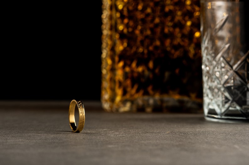 May include: A gold ring with a textured surface and engraved lettering. The ring is lying on a gray surface.