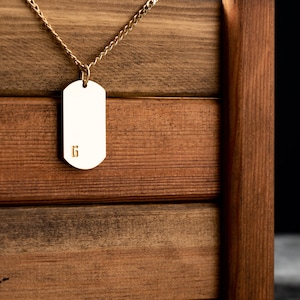 May include: A gold chain necklace with a blank gold dog tag pendant. The pendant has the number "6" engraved on it. The necklace is hanging from a wooden box.