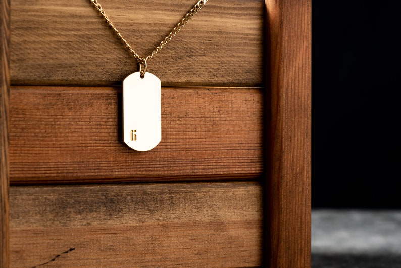 May include: A gold chain necklace with a blank gold dog tag pendant. The pendant has the number "6" engraved on it. The necklace is hanging from a wooden box.