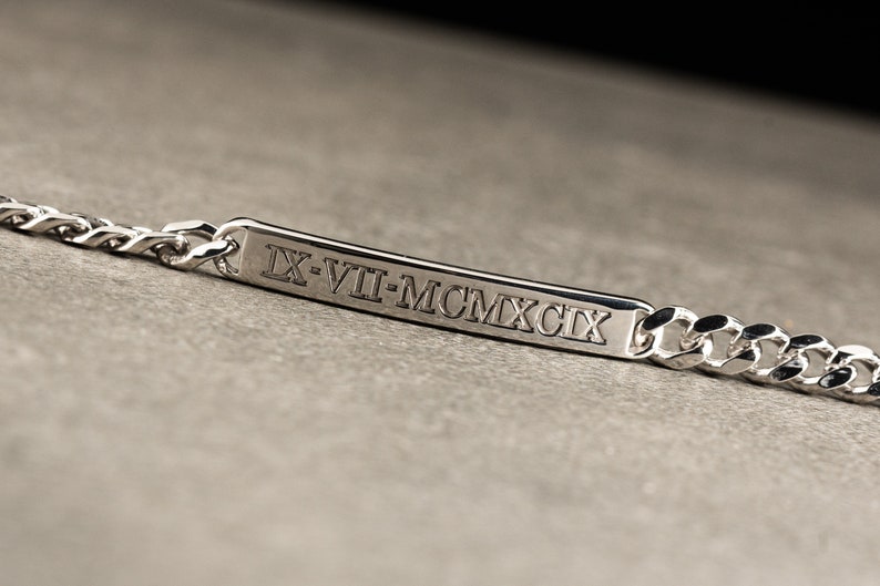 K&ouml;nnte beinhalten: Silberarmband mit einer rechteckigen Plakette, die mit dem r&ouml;mischen Datumszeichen "IX-VII-MCMXCIX" graviert ist.