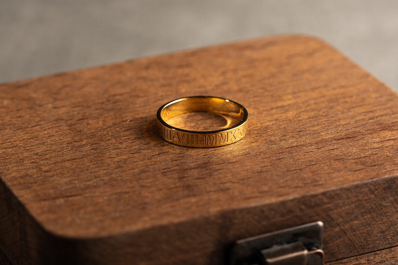 May include: A gold ring with Roman numerals engraved around the band. The ring is sitting in a wooden box.