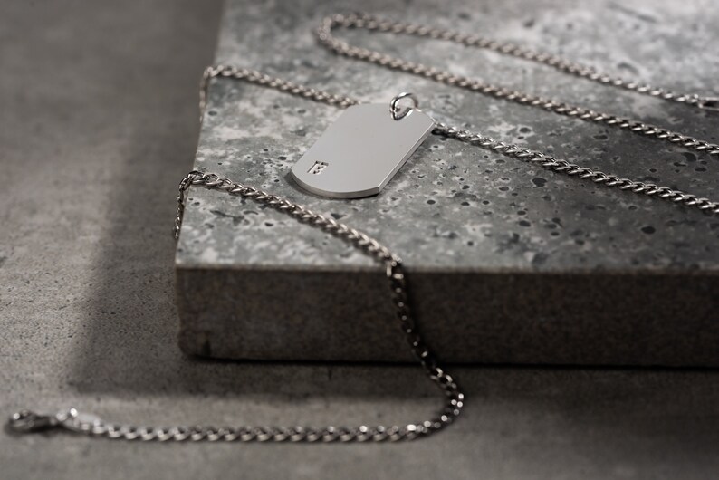 May include: A silver chain necklace with a silver dog tag pendant. The dog tag has a small inscription on it.