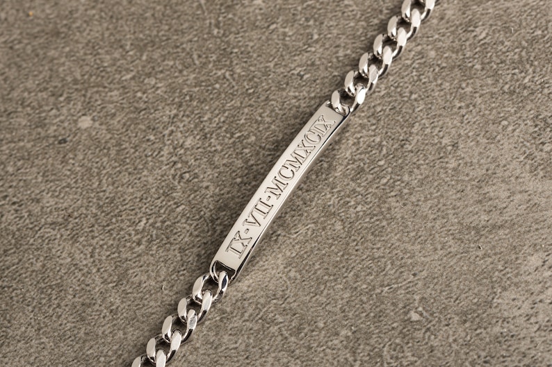 K&ouml;nnte beinhalten: Ein silbernes Kettenarmband mit einem rechteckigen Anh&auml;nger, der mit dem r&ouml;mischen Datumszeichen "IX-VII-MCMXCIX" graviert ist.