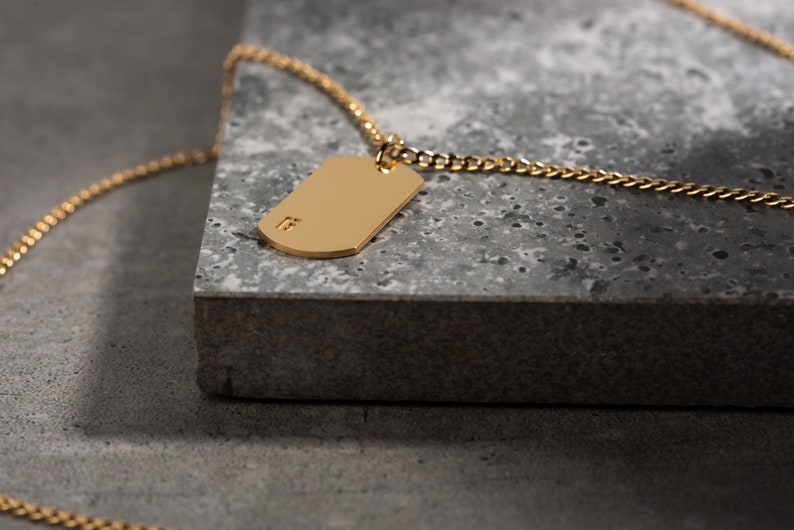 May include: A gold chain necklace with a gold dog tag pendant. The dog tag has a small inscription on it.