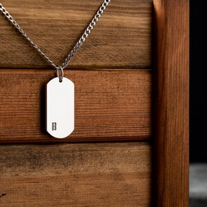 May include: A silver chain necklace with a silver dog tag pendant. The dog tag has the letter "B" engraved on it. The necklace is hanging on a wooden surface.