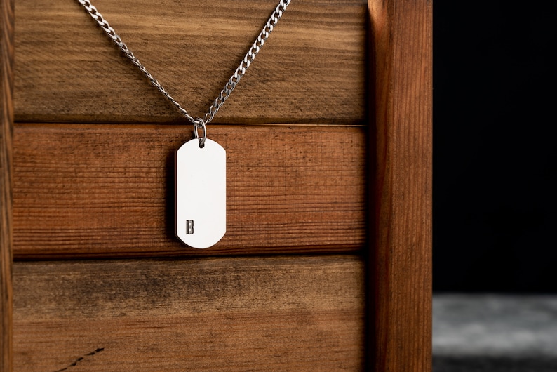 May include: A silver chain necklace with a silver dog tag pendant. The dog tag has the letter "B" engraved on it. The necklace is hanging on a wooden surface.