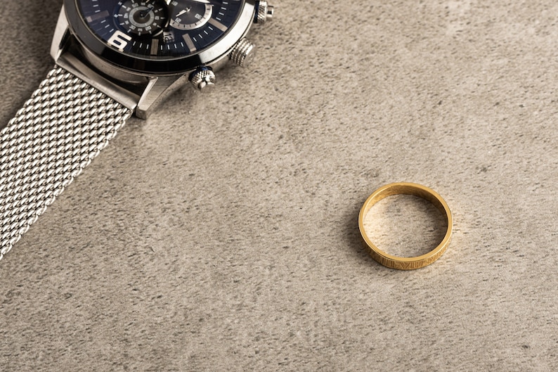 May include: A silver watch with a black face and a mesh band, and a gold ring on a gray background.