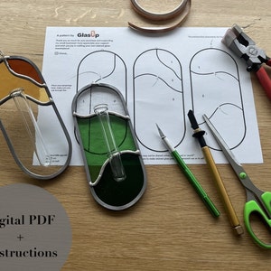 Puede incluir: Un patrón de GlasUp para proyectos de vitrales. La imagen muestra dos jarrones de vitrales terminados, uno con un diseño amarillo y marrón y el otro con un diseño verde. La imagen también muestra las herramientas y los materiales necesarios para el proyecto, incluyendo cinta de cobre, un cortador de vidrio, una amoladora de vidrio y un par de tijeras.