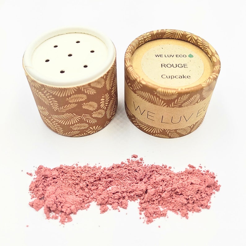 May include: Two brown cardboard containers with a leafy pattern and a white lid with holes. The container on the right has a label that reads "WE LUV ECO ROUGE Cupcake". A pile of pink powder is spilled out in front of the containers.