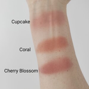 May include: Three swatches of blush on a person's arm. The swatches are labeled "Cupcake", "Coral", and "Cherry Blossom".