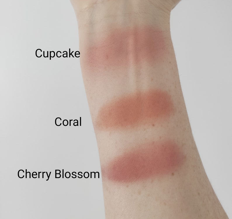 May include: Three swatches of blush on a person's arm. The swatches are labeled "Cupcake", "Coral", and "Cherry Blossom".