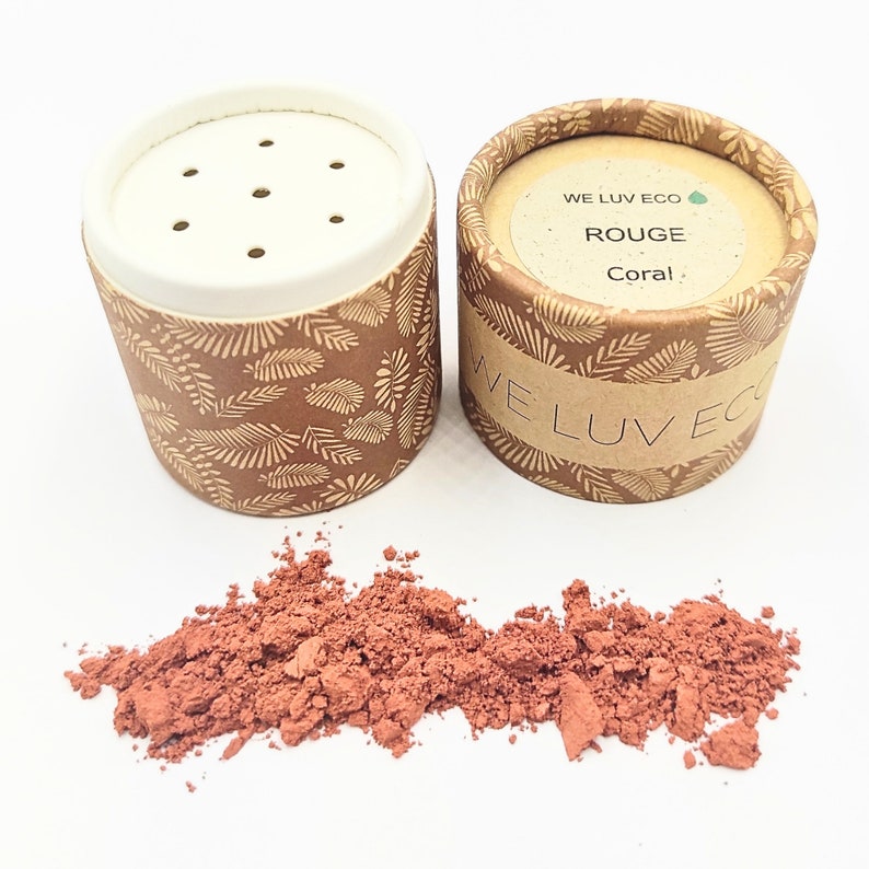 May include: Two cardboard containers with a leafy pattern and a white lid with holes. The container on the right has the text "WE LUV ECO" and "ROUGE Coral". A pile of red powder is in front of the containers.