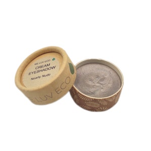 May include: A small, round container of cream eyeshadow in the shade "Nearly Nude". The container is made of cardboard and has a brown and white pattern. The lid of the container is brown and has the text "WE LUV ECO" and "CREAM EYESHADOW" printed on it.