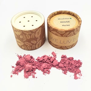 May include: A small, round, cardboard container with a white lid and a brown and gold leaf pattern. The container is labeled "WE LUV ECO ROUGE Merlot". The container is open and a pile of pink powder is spilling out.