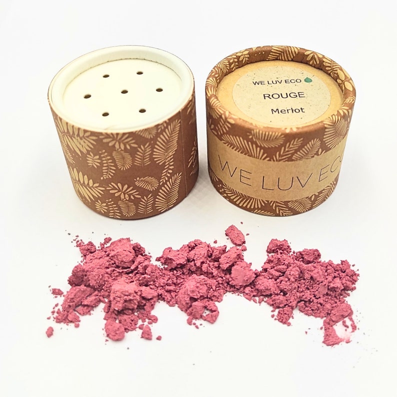 May include: A small, round, cardboard container with a white lid and a brown and gold leaf pattern. The container is labeled "WE LUV ECO ROUGE Merlot". The container is open and a pile of pink powder is spilling out.