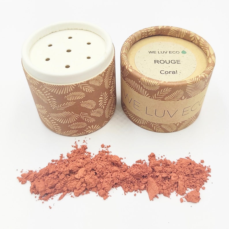 May include: A small cardboard container with a white lid and a leaf pattern. The container is labeled "WE LUV ECO" and "ROUGE Coral". The container is open and a pile of red powder is spilling out.