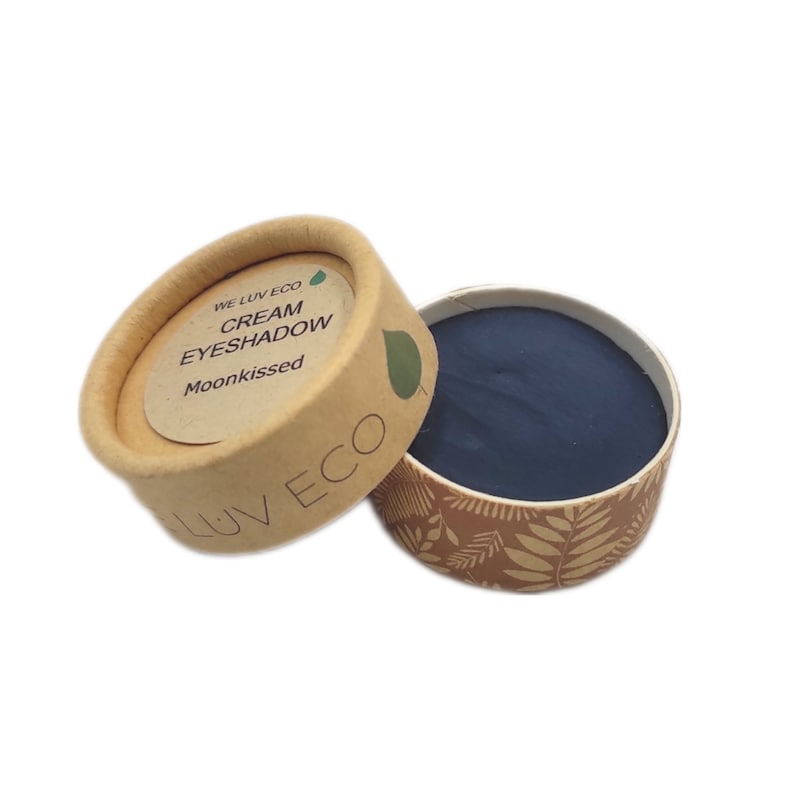 May include: A small, round, cardboard container with a lid. The container is brown with a floral pattern and the lid has the text "WE LUV ECO CREAM EYESHADOW Moonkissed" on a white label. The container is open and shows a small, round, dark blue cream eyeshadow inside.