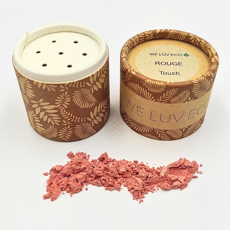 May include: A small, round container of loose red blush powder. The container is made of cardboard and has a brown and gold floral pattern. The container has a white lid with holes for dispensing the powder. The label on the container reads "WE LUV ECO ROUGE Touch".