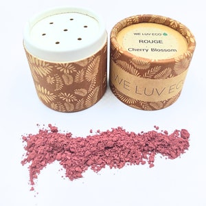 May include: Two small cardboard containers with a brown and gold leaf pattern. The container on the right has the text "WE LUV ECO" and "ROUGE Cherry Blossom". The container on the left is open and shows a pink powder inside.