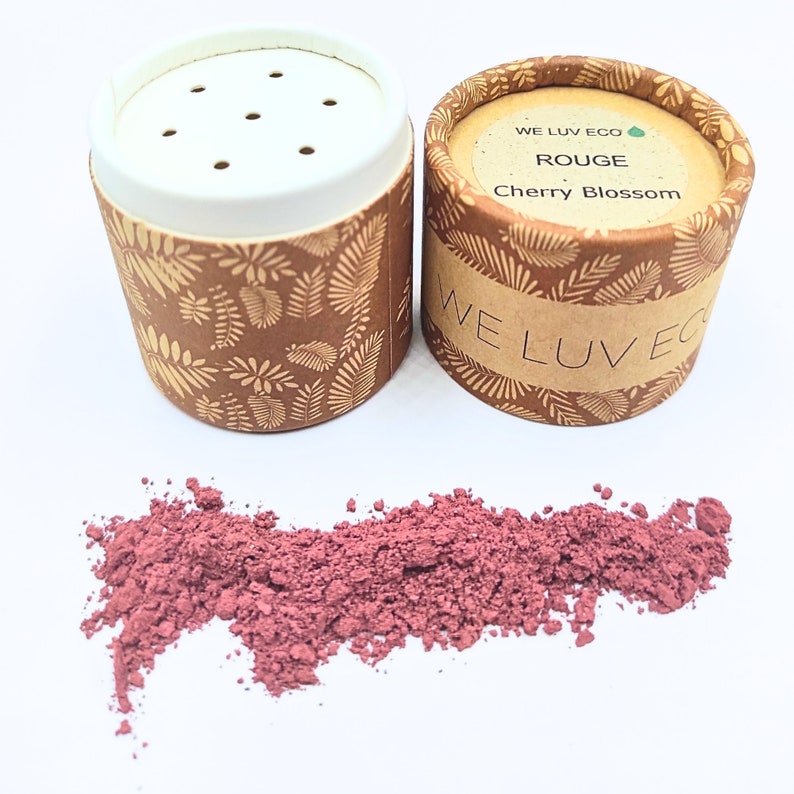 May include: Two small cardboard containers with a brown and gold leaf pattern. The container on the right has the text "WE LUV ECO" and "ROUGE Cherry Blossom". The container on the left is open and shows a pink powder inside.