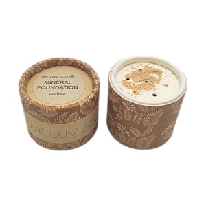 Puede incluir: Dos envases cilíndricos de base mineral. Uno está cerrado, mostrando el texto "WE LUV ECO MINERAL FOUNDATION Vanilla". El otro está abierto, revelando el polvo. Ambos envases son marrones con un patrón de hojas.