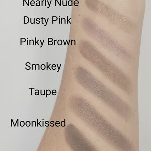 May include: Swatch of eyeshadow colors on a person's arm. The colors are labeled from lightest to darkest: Golden Beige, Nearly Nude, Dusty Pink, Pinky Brown, Smokey, Taupe, and Moonkissed.