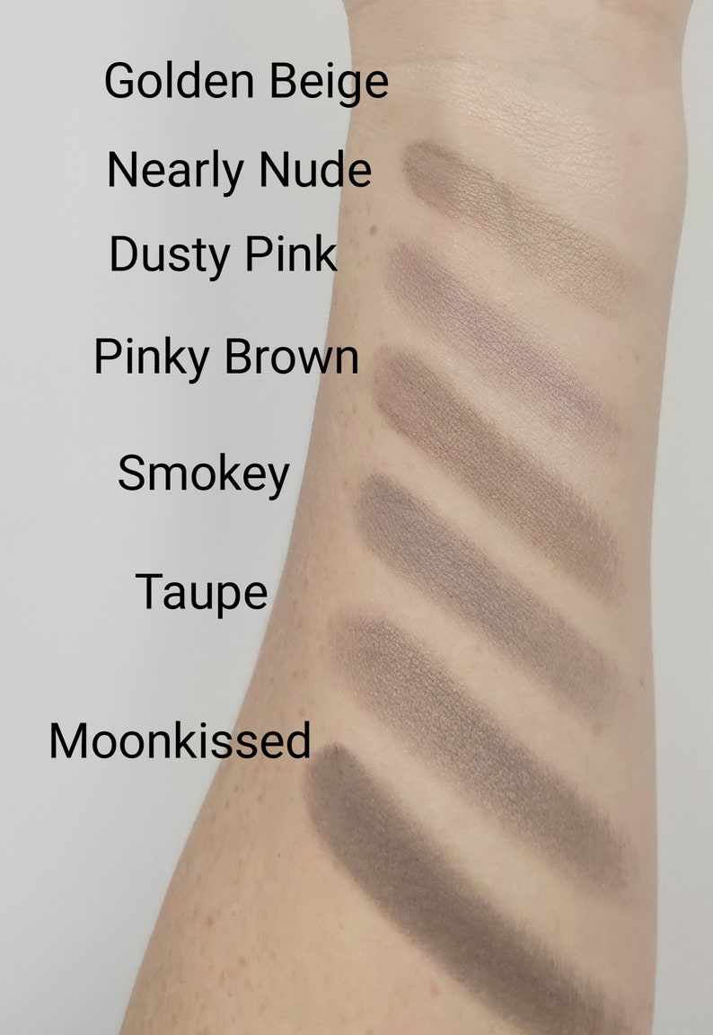 May include: Swatch of eyeshadow colors on a person's arm. The colors are labeled from lightest to darkest: Golden Beige, Nearly Nude, Dusty Pink, Pinky Brown, Smokey, Taupe, and Moonkissed.