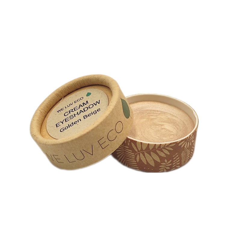 May include: A small, round container of cream eyeshadow in the shade Golden Beige. The container is made of cardboard and has a brown and white leaf pattern. The lid of the container is labeled "WE LUV ECO CREAM EYESHADOW Golden Beige".