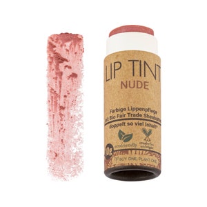 Puede incluir: Un tubo de cartón marrón con un tinte labial en color nude. El tubo tiene el texto "LIP TINT NUDE" impreso en él. El tubo también tiene el texto "Farbige Lippenpflege mit Bio Fair Trade Sheabutter doppelt so viel Inhalt" impreso en él. El tubo tiene una hoja verde y un símbolo de corazón con el texto "ecofriendly crueltyfree andvegan" impreso en él. El tubo tiene el texto "BUY ONE PLANT ONE" impreso en él.