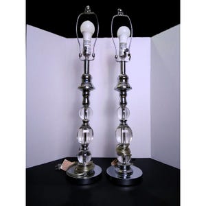 May include: A pair of tall table lamps featuring a polished chrome finish, each with a central column composed of three stacked clear glass spheres. The lamps include a silver-toned metal base, a harp for a shade, and a white LED light bulb.
