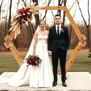 May include: A couple stands in front of a wooden hexagonal arch decorated with red flowers and pampas grass. The bride wears a white strapless gown and veil, holding a bouquet of red roses. The groom wears a black suit with a red boutonniere. The setting is outdoors.