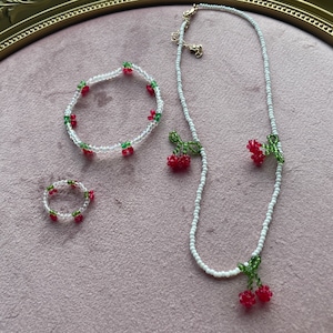 May include: A beaded jewelry set featuring a necklace, bracelet, and ring. The jewelry is adorned with red cherry and green leaf accents. The necklace and bracelet are strung with white beads. The ring is a small circle of white beads with cherry and leaf accents.