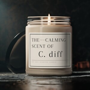 May include: A beige soy wax candle in a glass jar with a black lid. The candle label reads "THE CALMING SCENT OF C. diff" and "Soy Wax Scented Candle".