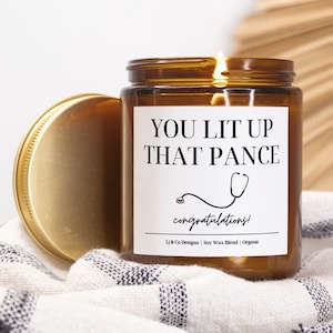 May include: A lit candle in a brown glass jar with a gold lid. The label reads "YOU LIT UP THAT PANCE" with a stethoscope graphic and "congratulations!". The candle is on a black and white striped cloth.